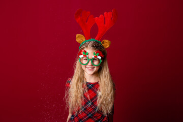 Wall Mural - cute little girl in Christmas glasses blows snow from palms in the studio on a red background. Christmas concept, text space