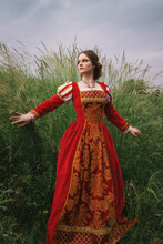 Beautiful Young Woman In A Long Red Medieval Dress Is Standing In The Grass In The Field Under The Cloudy Sky, Fantasy Princess