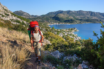 A sporty man with a large backpack climbs a narrow path overlooking the Mediterranean Sea. Turkey