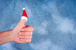 Hand with red Santa's hat on thumb up, blue concrete background. Merry Christmas, like sign, New Year, funny card concept. Close-up, copy space