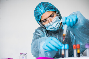 Blood test tube with label of Covid-19 also known as coronavirus or novel corona in scientist hand in laboratory. Female scientist is wearing highly protective suit, gloves, mask and glasses.