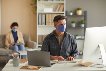 Wall Mural - businessman in protective mask working on computer at his table with his colleague in the background