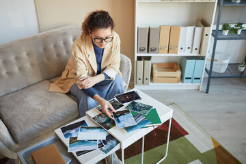 Wall Mural - young woman in eyeglasses sitting on sofa at the table and choosing the best photos for design proje