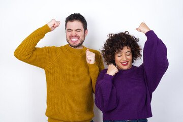 Sticker - Attractive Young couple wearing knitted sweater standing against white wall celebrating a victory punching the air with his fists and a beaming toothy smile.