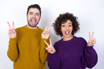 Wall Mural - Young couple wearing knitted sweater standing against white wall with optimistic smile, showing peace or victory gesture with both hands, looking friendly. V sign.