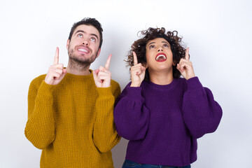 Wall Mural - Successful friendly looking Young couple wearing knitted sweater standing against white wall exclaiming excitedly, pointing both index fingers up, indicating something.