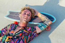 Funky Young Male Skateboarder In Trendy Colorful Shirt Resting Lying On Skate In Skatepark