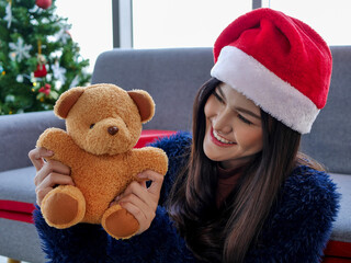  Asian attractive smiling happy woman wearing red Santa hat and teddy bear as gift with Christmas tree during holiday festival celebration at home new normal