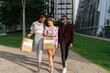 © Luis Manuel Munoz/ADDICTIVE STOCK - Full length of group of modern millennial multiethnic friends in fashionable outfits holding shopping bags while standing together against contemporary city buildings with glass walls