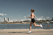 © Sergey Mironov/ADDICTIVE STOCK - Side view full body of energetic motivated fit female in activewear with fitness tracker jogging on paved embankment near river in city