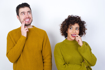 Sticker - Young couple wearing knitted sweater standing against white wall with thoughtful expression, looks to the camera, keeps hand near face, bitting a finger thinks about something pleasant.