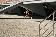 Side View Of Determined Female Athlete Running Fast During Outdoor Workout Near Stairways Of Stadium