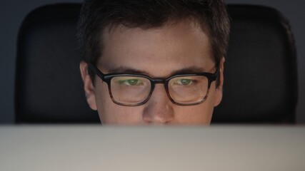Closeup of man eyes in eyeglasses in front of the computer in home office on background of grey wall. Young man sitting at home working alone at night