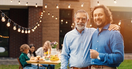 Portrait of handsome Caucasian two men, old and young, standing in hugs at back yard of house. Family dinner outdoor on background. Senior father with adult son embracing. Dad pensioner. Generations.