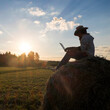 © Aliyev Alexei Sergeevich/Blend Images - Mari man sitting on hay bale at sunset using laptop