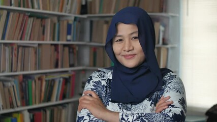 Wall Mural - Portrait of cheerful Asian muslim female librarian wearing hijab, smiling crossed arms confidence gesture against book shell in library