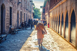 Fototapeta Uliczki - Turkish woman walking down cobblestone narrow street with benches near old traditional buildings in Istanbul, crowd of men background, view from back against sun, Turkey