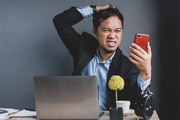 Stressed young businessman in modern workplace looking at mobile phone and scratches his head looks like it's getting bad news or negative news with a disappointed expression on grey background