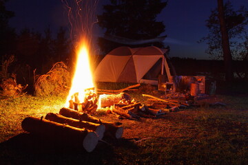 Beautiful campfire with bright orange flame light, firewood and camping tent on campsite at calm summer night, romantic lifestyle outdoor tourism vacation on nature landscape in Russia on empty glade