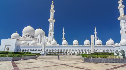 Wall Mural - Sheikh Zayed Grand Mosque timelapse hyperlapse in Abu Dhabi, the capital city of United Arab Emirates. Side view with domes and minarets. Trees on foreground. Blue sky at sunny day