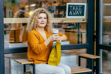 Wall Mural - Blond curly female customer in yellow knitted cardigan, sitting outdoor near glass window of store or zero waste shop, on bench, drinking coffee from reusable silicone cup at warm aututmn day.