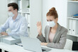 © pressmaster - Young businesswoman in protective mask waving hand to someone on laptop display