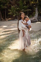 Wall Mural - A young tall, long-haired brunette, a fair-haired girl, a mother, is walking along the azure sea and carries a small, fair-haired, happy son in her arms.white cotton clothes. boho dress.