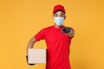 Wall Mural - Delivery employee african man in red cap blank print t-shirt face mask gloves uniform work courier service on quarantine covid-19 virus concept hold cardboard box isolated on yellow background studio.