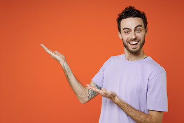 Wall Mural - Smiling cheerful young bearded man 20s wearing casual violet t-shirt standing pointing hands aside on mock up copy space looking camera isolated on bright orange color wall background studio portrait.