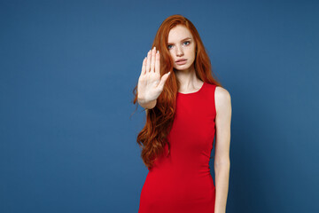Wall Mural - Displeased dissatisfied young redhead woman 20s wearing bright red elegant evening dress standing showing stop gesture with palm looking camera isolated on blue color wall background studio portrait.
