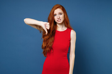 Displeased dissatisfied confused young redhead woman 20s wearing bright red elegant evening dress standing showing thumb down looking camera isolated on blue color wall background studio portrait.