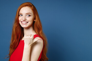 Wall Mural - Side view of smiling pretty young redhead woman 20s wearing bright red elegant evening dress standing pointing thumb aside on mock up copy space isolated on blue color wall background studio portrait.