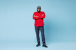 © ViDi Studio - Full length of smiling handsome young bearded african american man in casual red shirt cap eyeglasses standing holding hands crossed looking camera isolated on pastel blue background studio portrait.