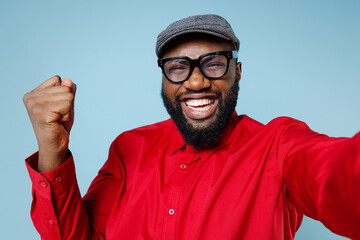 Close up of joyful young bearded african american man 20s in red shirt eyeglasses cap doing selfie shot on mobile phone doing winner gesture isolated on pastel blue color background studio portrait.