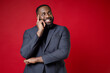 © ViDi Studio - Smiling funny pensive young african american business man 20s wearing classic jacket suit standing put hand prop up on chin looking aside isolated on bright red color wall background studio portrait.