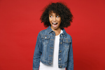 Excited amazed little african american kid girl 12-13 years old wearing casual denim jacket looking aside isolated on bright red color background children studio portrait. Childhood lifestyle concept.