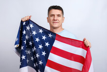 Attractive Middle-aged Man In Casual T-shirt Holds And Showing In His Arms American Flag And Looking At Camera On Grey Background. 