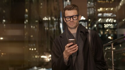 Man in eyeglasses with phone in hand on background of building with lights on. Handheld night shot of caucasian man texting in phone near business building with cars