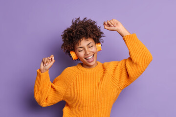Canvas Print - Positive dark skinned young woman dances carefree wears orange jumper moves with rhythm of music uses stereo headphones isolated over purple background. People hobby and entertainment concept