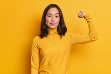 Serious Self Confident Brunette Young Asian Woman Raises Hand Up And Shows Demonstrates Strength Looks Like Strong Powerful Bodybuilder Dressed In Casual Turtleneck Isolated Over Yellow Background