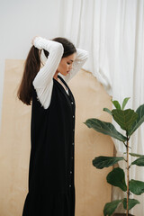 Side view of a young beautiful sensual woman putting her long hair in a tail. She's wearing black dress over white long sleeve shirt.