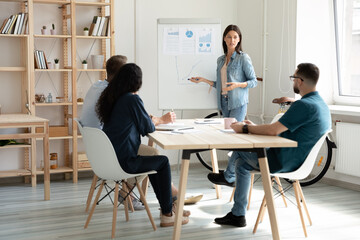 Wall Mural - Young Caucasian female speaker or coach make flip chart presentation for diverse colleagues at office meeting. Successful woman trainer present project on whiteboard for employees at briefing.