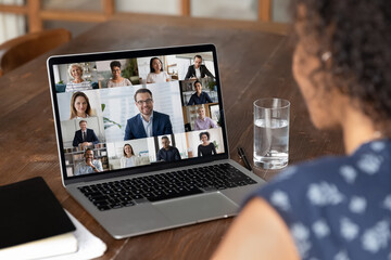 Wall Mural - Back shoulder view young african american woman holding video conference web camera call with diverse multiracial colleagues and leader, working distantly from home, discussing project ideas online.