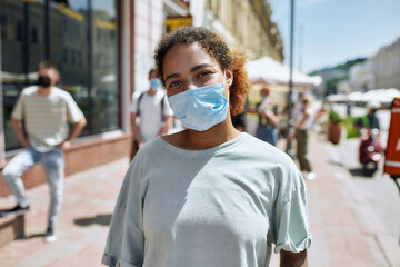 Wall Mural - Portrait of young african american woman wearing mask looking at camera. People collecting their orders from the pickup point during coronavirus lockdown in the background