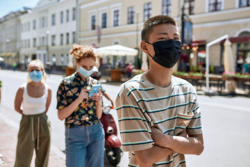 Wall Mural - Young asian guy wearing mask waiting in line, respecting social distancing to enter takeout restaurant or to collect purchases from the pickup point during coronavirus lockdown