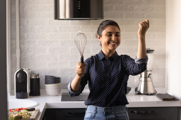 Singer in heart. Enthusiastic young indian woman enjoy preparing food at modern home kitchen. Funny young hindu female take break in cooking listen to music dance sing with microphone of egg beater