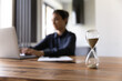 © fizkes - Busy young mixed race woman working studying by laptop on blurred background doing urgent job taking exam. Focus on close up sand glass posed on desk at home office. Measuring time. Deadline is coming
