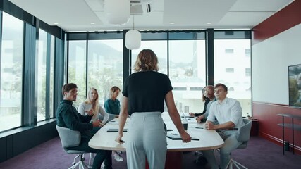 Wall Mural - Rear view of a businesswoman walking in the board room with staff sitting around table.  Office manager arriving in the conference room with her team settling down around the table.
