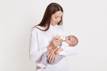 Wall Mural - Mother feeding her baby from feeding bottle, dark haired female wearing sweater hold baby in hands and looking at her kid, posing isolated over white background.