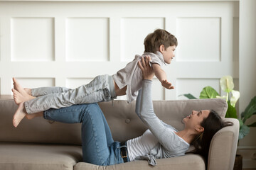 Wall Mural - Playful young Caucasian mother lying on sofa in living room play with excited small preschooler son. Happy mom or nanny hold in hands imitate plane with little boy child, enjoy family weekend at home.